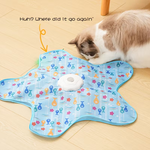 Cat playing with a fish-patterned mat on a wooden floor