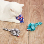 White cat playing with two colorful cat toys on a wooden floor.