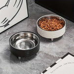 Two pet bowls on a marble surface, one black and one white with a silver interior, containing dry cat food.